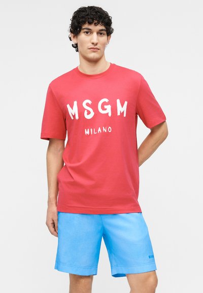 Νέος άνδρας με σγουρά σκούρα μαλλιά, φόρεμα ένα κόκκινο T-shirt MSGM Milano και γαλάζια σορτς, στέκεται με το ένα χέρι πίσω από την πλάτη του.