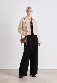 Giacca in denim beige corta sopra una maglietta nera, abbinata a pantaloni neri a gamba larga. Una piccola borsa rossa con tracolla a catena completa il look.