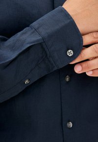 Camicia a maniche lunghe blu scuro realizzata in tessuto strutturato. Presenta polsini con bottone e bottoni coordinati lungo la parte anteriore. Finitura liscia.
