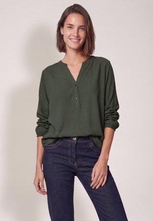 Mujer sonriendo, vistiendo una blusa verde oscuro de manga larga con botones y vaqueros azul oscuro, de pie frente a un fondo claro y liso.