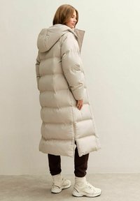 Abrigo acolchado largo beige con capucha, con un diseño acolchado, cremallera frontal y aberturas laterales, combinado con pantalones oscuros y zapatillas claras.