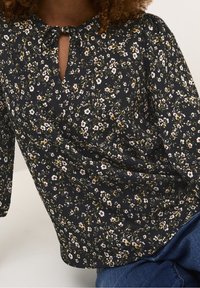 Sort bluse med blomsterprint i pink og grøn, med nøglehuludskæring og sløjfedetalje samt lange, puffede ærmer. Tekstureret stof.