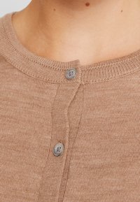 Světle hnědý pletený cardigan s ribovanou strukturou, který má kulatý výstřih a dva šedé knoflíky vpředu pro zapínání.