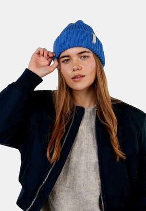 Blue ribbed knit beanie with a folded cuff and a light tan label. Model wearing a black bomber jacket over a beige sweater.