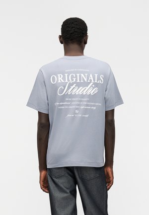 Persona que lleva una camiseta gris con texto blanco "ORIGINALS Studio" y texto adicional más pequeño en la espalda, combinada con jeans oscuros.