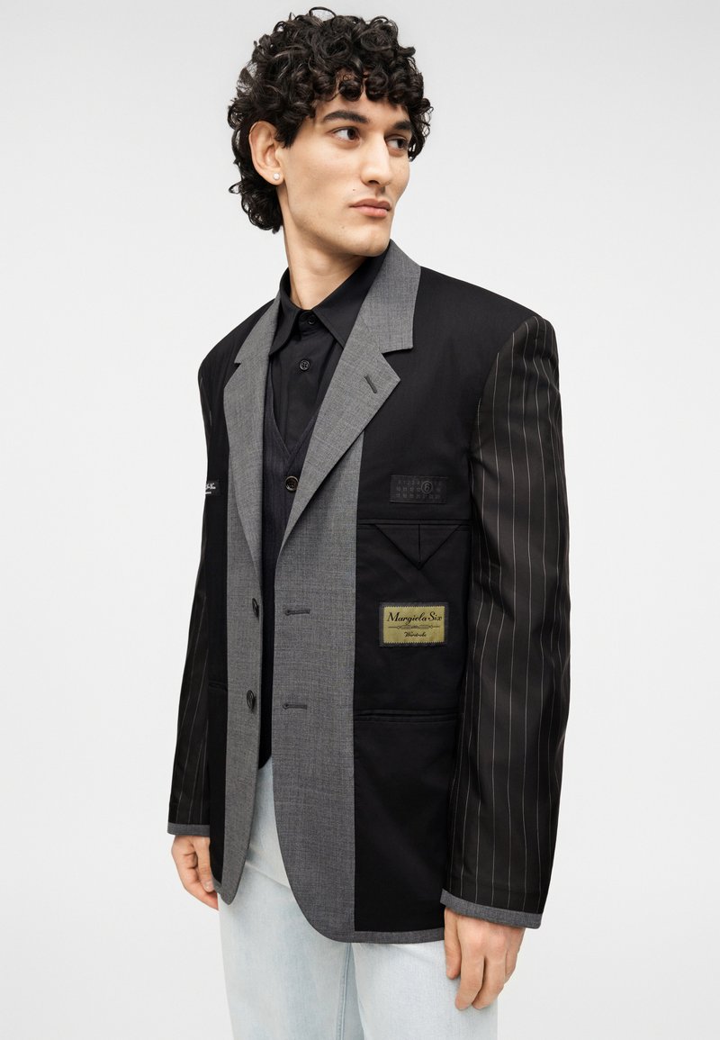 Blazer patchwork noir et gris avec un mélange de tissus unis et à fines rayures, doté d'une étiquette sur la poche poitrine et d'une coupe ajustée.