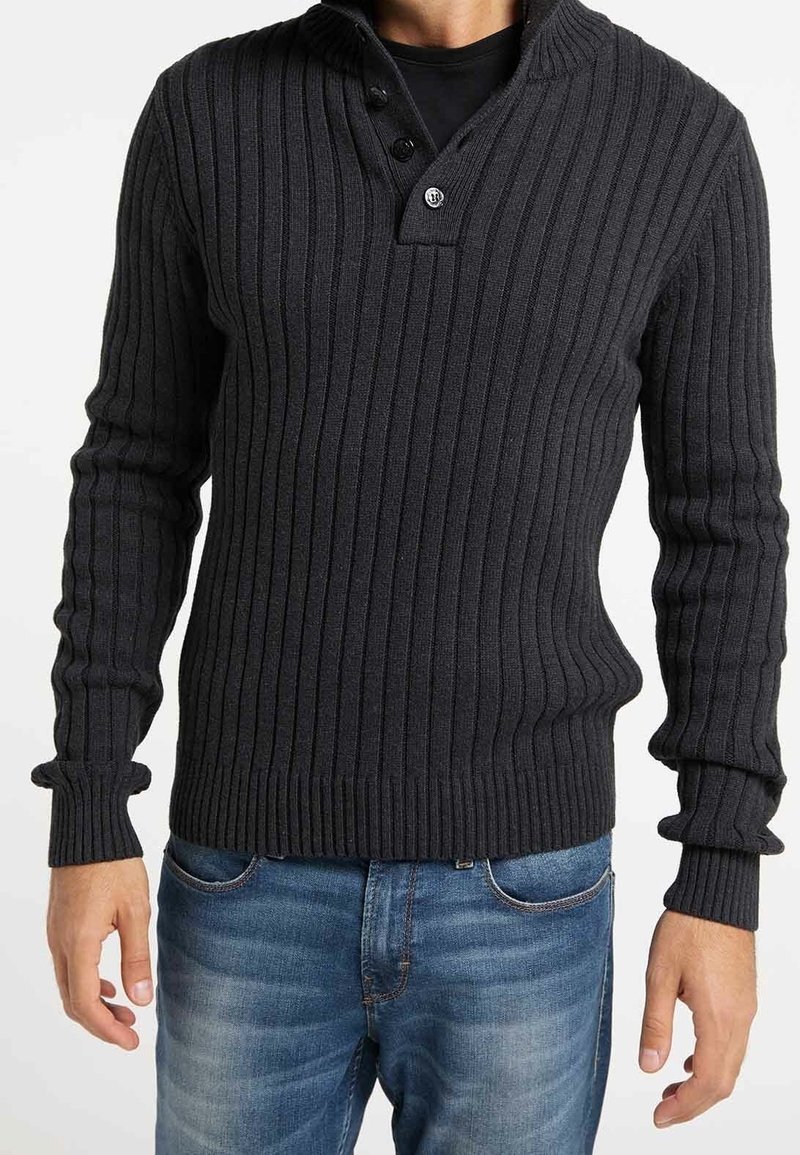 Homme portant un pull en maille côtelée gris foncé avec col boutonné et un jean en denim bleu, debout devant un fond blanc.