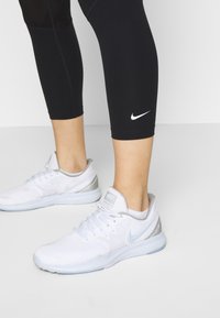 Białe buty sportowe Nike z jasnoszarymi akcentami i teksturowaną siateczką, zestawione z czarnymi legginsami 3/4 z białym logo Nike.