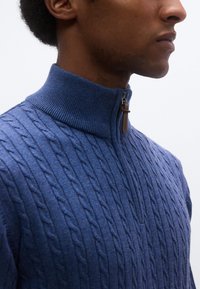 Μπλε πλεκτό πουλόβερ με ψηλό ριμπ γιακά, με υφασμάτινο μοτίβο καλωδίου και φερμουάρ με δερμάτινη ταινία έλξης.