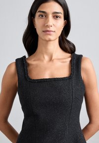 Kobieta z długimi ciemnymi włosami, ubrana w czarną, bezrękawnikową sukienkę o fakturowanym materiale z kwadratowym dekoltem i koralikowym wykończeniem, na jednolitym szarym tle.