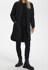 Schwarze, gesteppte lange Jacke mit Reißverschluss, seitlichen Taschen und gerippten Bündchen, kombiniert mit schwarzen, engen Jeans und Stiefeletten.