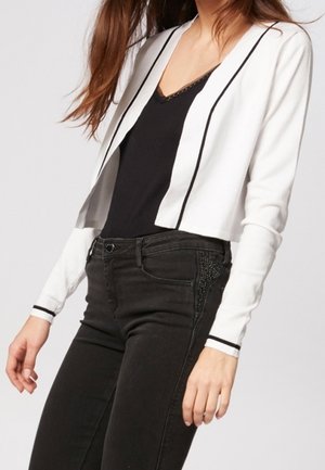 Frau trägt eine weiße, kurz geschnittene Strickjacke mit schwarzem Besatz über einem schwarzen Oberteil und hoch taillierten schwarzen Jeans mit verzierten Seitentaschen.