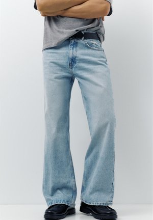 Person iført lyseblå vide jeans, sort bælte, grå skjorte og sorte loafers, stående med armene krydset mod en ensfarvet baggrund.