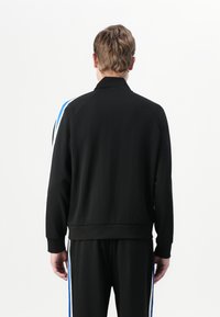 Veste de sport noire avec un col montant et des rayures bleues et blanches sur les épaules. Tissu léger et lisse avec une coupe décontractée.