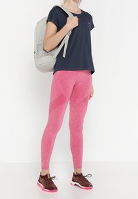 Žena v námořnickém tričku, růžových legínách, hnědých teniskách s růžovými podrážkami a nesoucí světle šedý batoh přes jedno rameno.