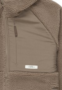 Giacca in pile di colore marrone chiaro con un'esterno testurizzato e un interno liscio. Presenta una tasca con zip e un'etichetta sul petto.