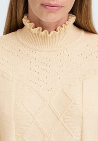 Pull en tricot beige clair avec un col montant à volants, présentant un motif texturé et de petits trous au niveau du décolleté, rehaussé de formes en losange.
