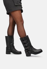 Botas de cuero negras hasta la rodilla con punta cuadrada, cremallera lateral y hebillas decorativas a lo largo del lado exterior. Diseño de tacón grueso para mayor estabilidad.