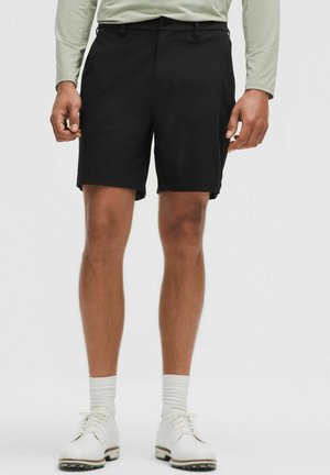 Mann trägt schwarze knielange Shorts, helles langärmeliges Hemd, weiße Socken und weiße Schnürschuhe, steht vor einfarbigem Hintergrund.