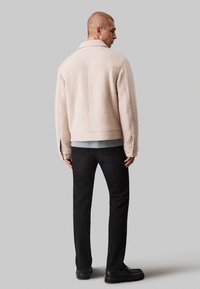 Veste en polaire beige avec un col, des coutures visibles et deux poches latérales, assortie à un jean noir et des chaussures noires. Vue de dos.