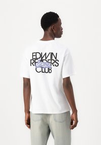 Hvit bomulls t-skjorte med stor svart tekst "EDWIN READERS CLUB" og et blått grafikk av sko på baksiden. Korte ermer og avslappet passform.