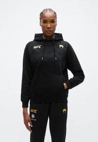 Sweat à capuche noir avec accents dorés, comprenant une poche avant, un design texturé sur les manches et le logo UFC sur la poitrine.