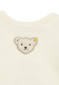 Cream-colored fabric with a round neckline; features an embroidered bear face with textured white fur, black eyes, and a yellow accent.