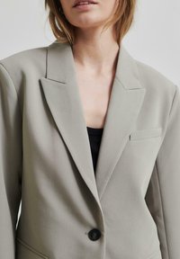 Lysegrå blazer med strukturerte fall, enkel knappelukking, og en liten brystlomme. Teksturet stoff, skreddersydd passform.