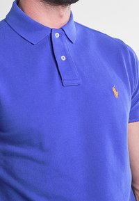 Homme portant un polo bleu roi avec deux boutons et un petit logo jaune brodé sur la poitrine gauche.