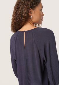 La blouse blu navy presenta un'apertura a chiave sul retro, un tessuto che si drappeggia morbido e maniche lunghe con una silhouette rilassata. Texture sottile e linee pulite.