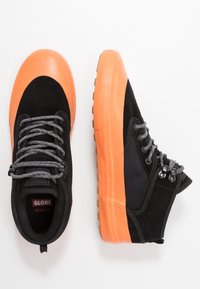 Schwarze und orangefarbene Sneaker mit Obermaterial aus Wildleder und Mesh, Gummisohle, strukturierten Akzenten und grauen Schnürsenkeln, mit einem Logo auf der Zunge.