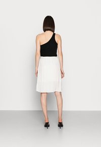 Femme de dos portant un top noir à une épaule, une jupe blanche jusqu'au genou et des sandales à talons noirs, se tenant contre un fond blanc.