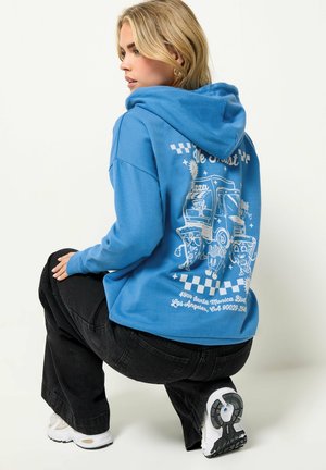 Blonde Frau hockt, trägt einen blauen Hoodie mit Cartoon-Pizza-Food-Truck-Design und schwarzen Hosen vor weißem Hintergrund.