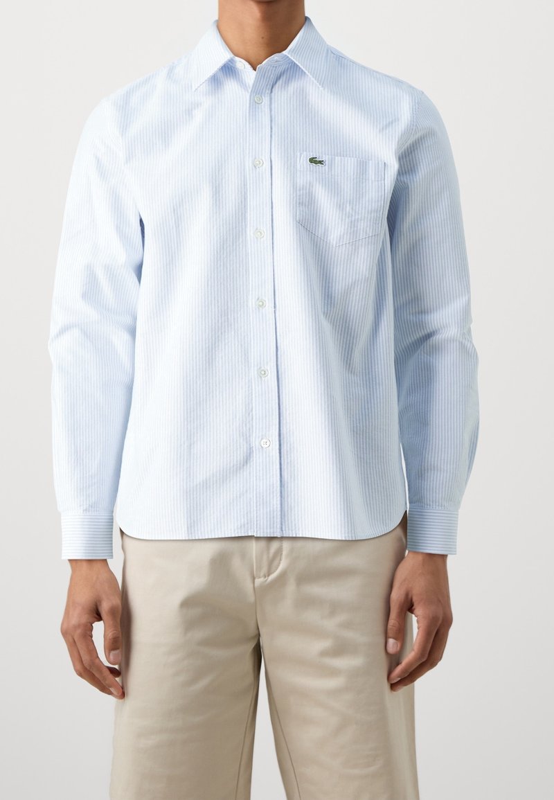 Jasnoniebieska, w paski, koszula z długim rękawem, z kieszonką na piersi i małym zielonym logo. Bawełniany materiał o gładkiej teksturze.