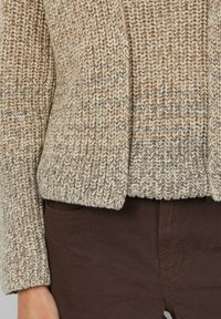 Strickpullover in Creme- und Taupe-Tönen, mit einem strukturierten Muster, weiten Ärmeln und einem cropped Design, kombiniert mit braunen Hosen.