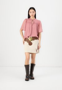 Rosa Bluse mit Polka-Dots und Puffärmeln, Schlüssel-ausschnitt. Beigefarbenen Rock mit einem Schlitz vorne und einem braunen Gürtel mit kreisförmigen Akzenten. Braune knielange Stiefel.