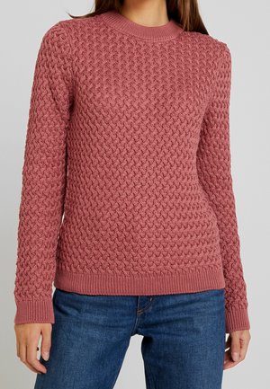 Femme portant un pull tricoté texturé rose poussiéreux à manches longues et col rond, associé à un jean bleu, sur un fond uni.