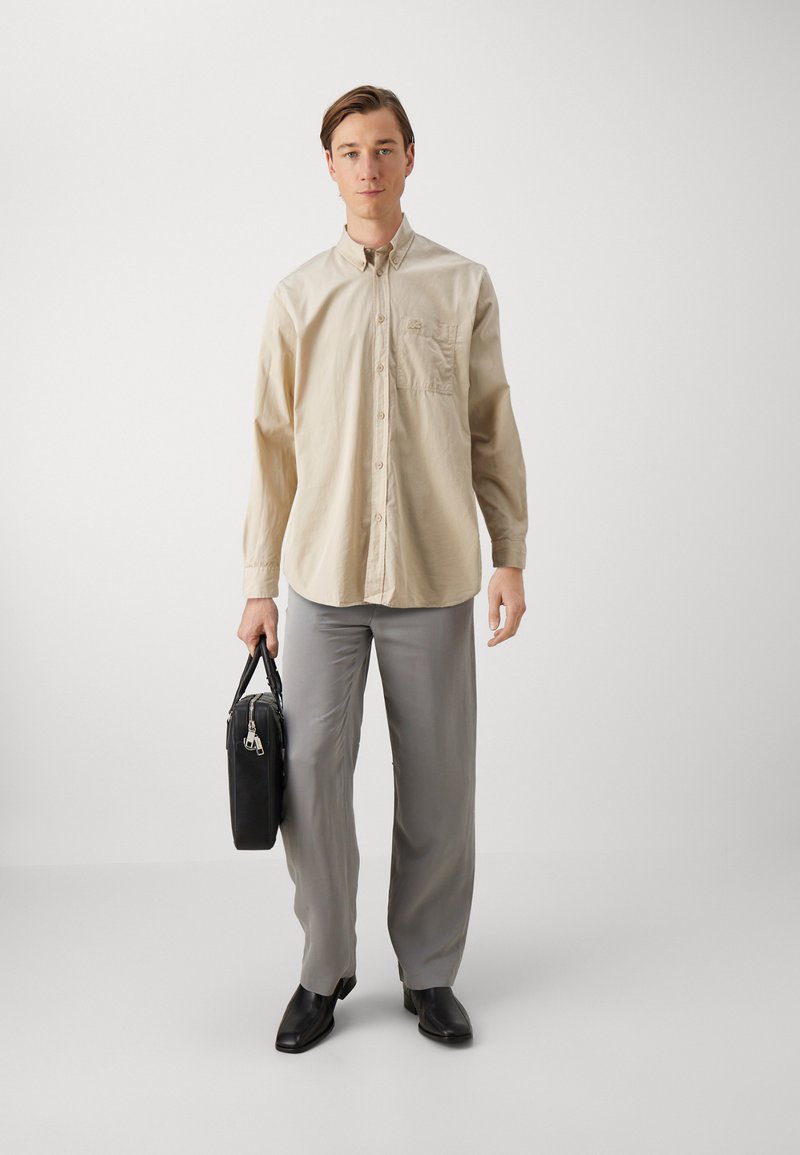 Beige overhemd met knopen en een borstzak, lichtgrijze broek, zwarte schoenen en een zwarte gestructureerde tas met een handvat aan de bovenkant.