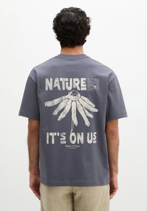 Mężczyzna w szarej koszulce z dużą białą grafiką kwiatową i napisem "NATURE IT'S ON US" na plecach na jednolitym tle.