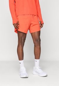 Ανδρικό μοντέλο φορώντας φωτεινές πορτοκαλί αθλητικές βερμούδες και αντίστοιχο μακρυμάνικο τοπ, λευκές κάλτσες Nike και λευκά αθλητικά παπούτσια, στέκεται σε ελαφρύ πάτωμα.