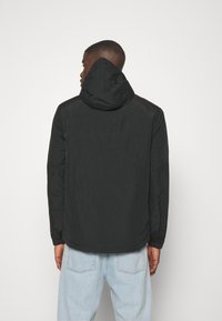 Giacca leggera nera con cappuccio, caratterizzata da una superficie liscia e un design minimalista. Cuciture visibili e vestibilità comoda. Abbinata a jeans azzurri chiari.
