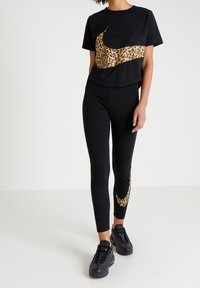 Persona che indossa un completo sportivo nero con un grande logo Nike a forma di leopardo sulla maglietta e un logo leopardo coordinato sui leggings, oltre a sneakers nere.