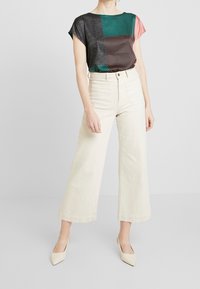 Mujer vistiendo una blusa de manga corta de bloques de color con múltiples patrones, pantalones de pierna ancha de talle alto en crema y tacones afilados en marfil.