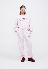 Ružový Nike sweatshirt s veľkým logom a zodpovedajúcimi teplákmi. Materiál je hladký; tepláky majú elastický pás a vrecká. Červené tenisky.