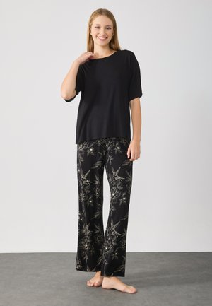 Ung kvinde smiler, iført en løs sort kortærmet bluse og sorte vide benbukser med beige blomster- og fuglemønstre, barfodet på grå gulv.