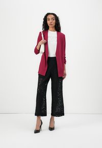 Blazer rouge, t-shirt blanc avec un graphisme, pantalon large en dentelle noire, et talons noirs. Le modèle porte un petit sac à main blanc.