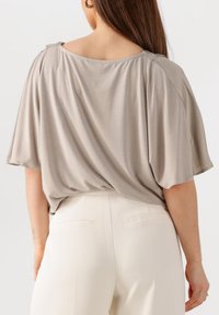 Loose-fitting, short-sleeved top in light gray fabric, featuring a wide neckline and gathered back detail. Paired with high-waisted white trousers.