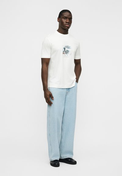 Λευκό κοντομάνικο μπλουζάκι με εκτύπωση σε ήπια χρώματα, συνδυασμένο με φαρδιά τζιν παντελόνια ανοιχτού μπλε και μαύρα παπούτσια.