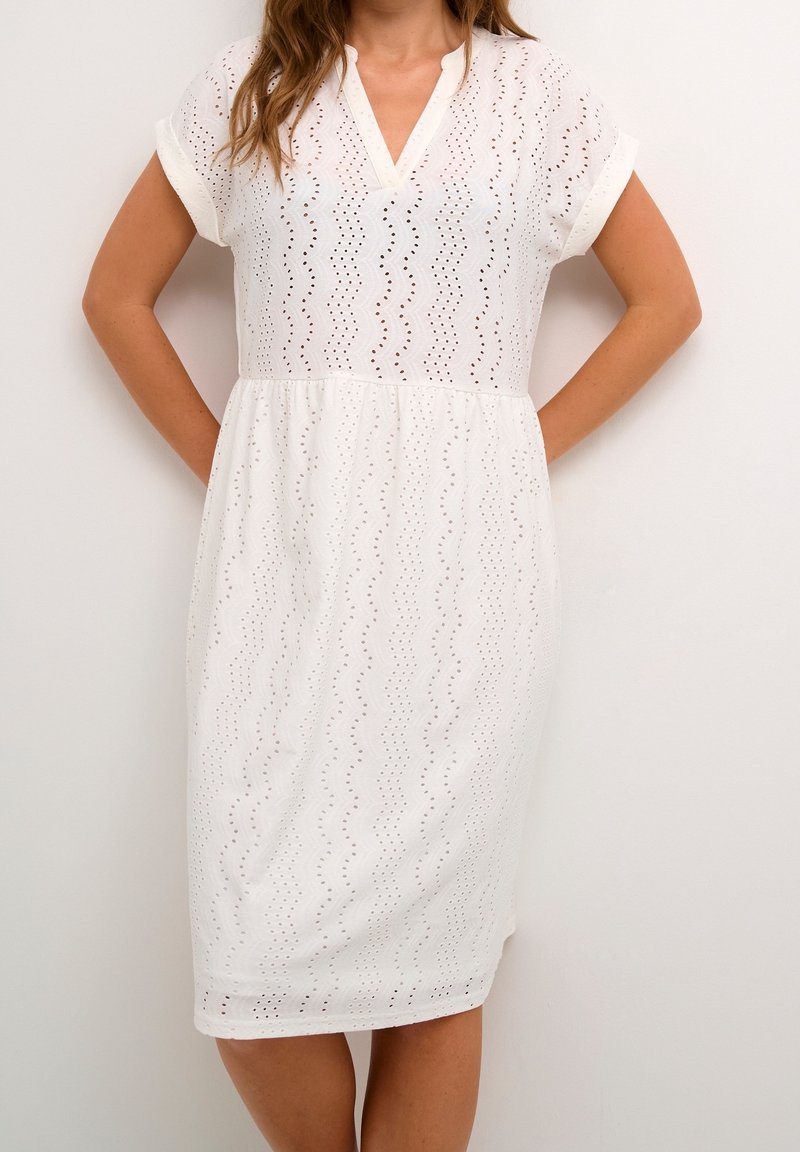 Femme portant une robe blanche à œillets, longueur genou, à manches courtes et col en V, debout les mains dans le dos.