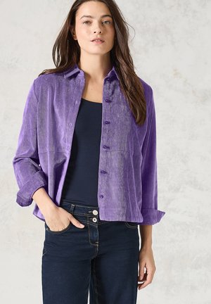 Lila Cord-Hemd mit Kragen, Knopfleiste und aufgerollten Ärmeln, kombiniert mit einem navyfarbenen Oberteil und dunklen Jeans.